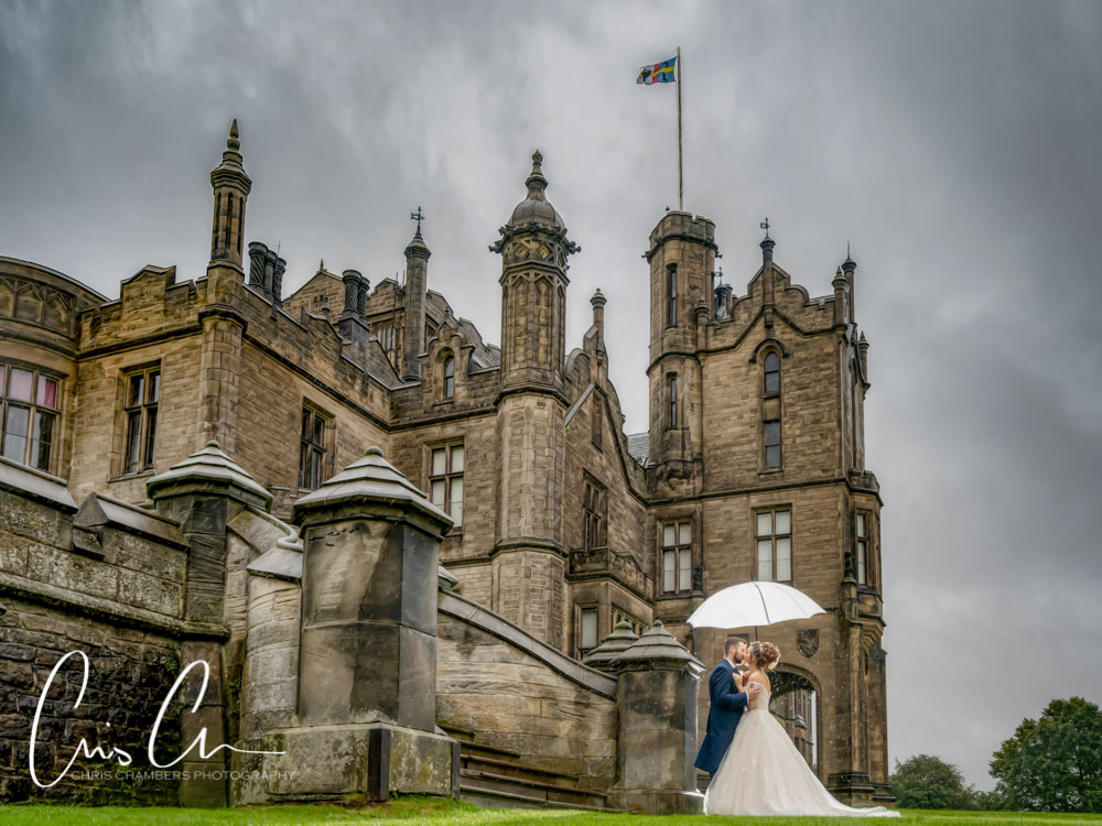 Wedding couple standing under an umbrella in front of a stately home.