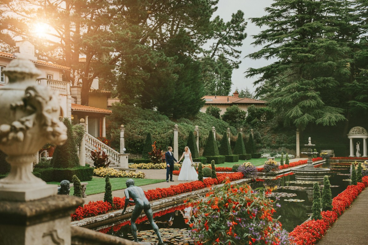 Couple On Their Wedding Day Standing In Manicured Gardens With Red Flowers
